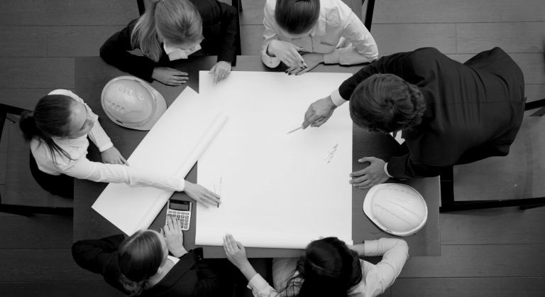 Team of six people gathered around a blueprint with hard hats on the table.
