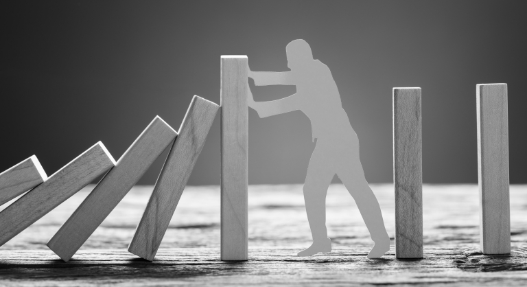 Paper man stopping wooden domino blocks on table against gray background.