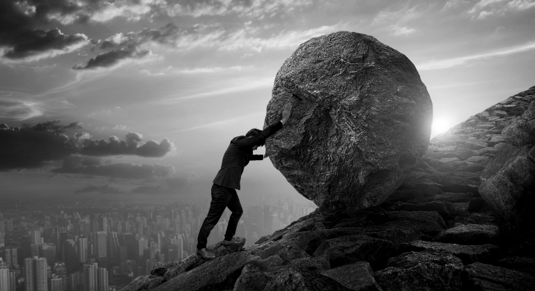 Business man pushing a large round stone up a steep stone hill.