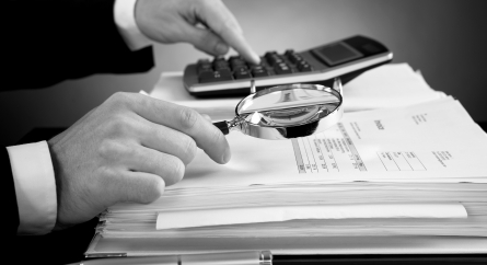 Person looking at a form on top of a stack of paper with a magnifying glass and calculator.