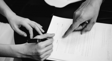 Close-up of 2 people's hands above a contract; one pointing where to sign and the other holding a pen about to sign the contract.
