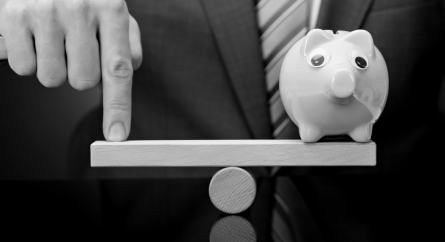 A piggy bank balancing on a scale with a person using a single finger to make it even.