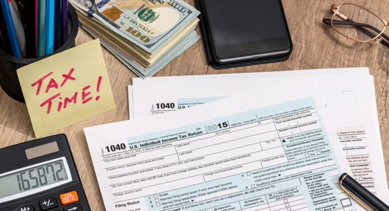 Tax forms on a desk with office supplies, a pile of money, a phone, glasses, and a sticky note that says "Tax Time!".