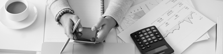 Woman at a table with her laptop, calculator, phone, notebook, charts, and a cup of coffee.