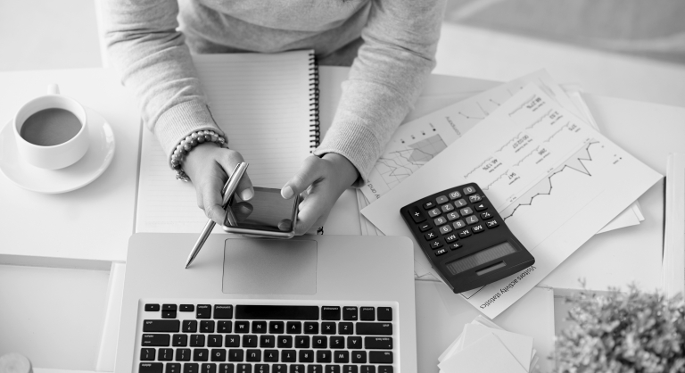 Woman at a table with her laptop, calculator, phone, notebook, charts, and a cup of coffee.