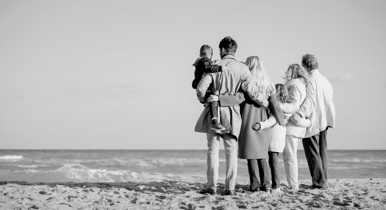 Shot from behind of a multigenerational family in coats on the beach.