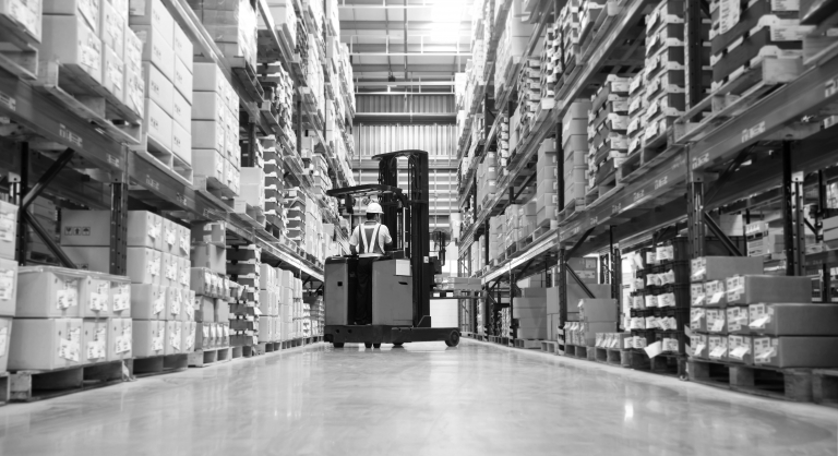 Worker in forklift-truck loading packed goods in huge distribution warehouse with high shelves.