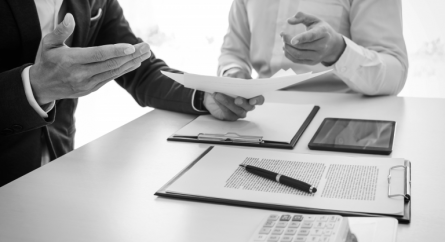 Two men sitting at a table with documents, a tablet, and a calculator discussing a document in one of the man’s hands.