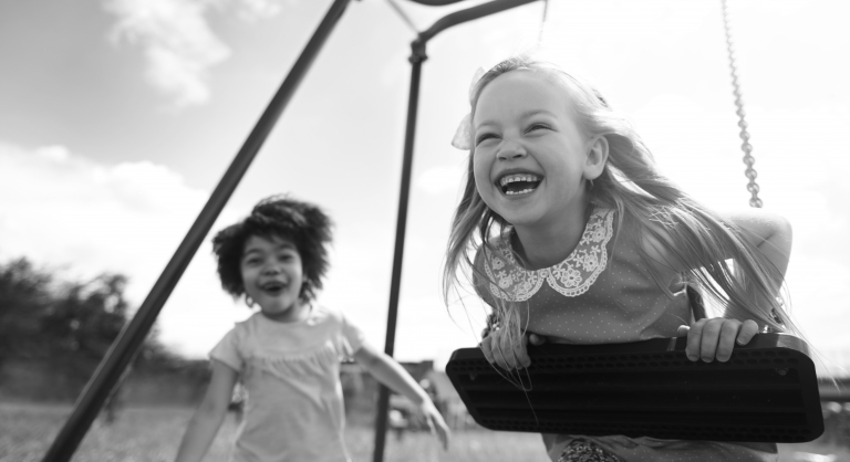 Two young kids laughing and playing together on a swing set.