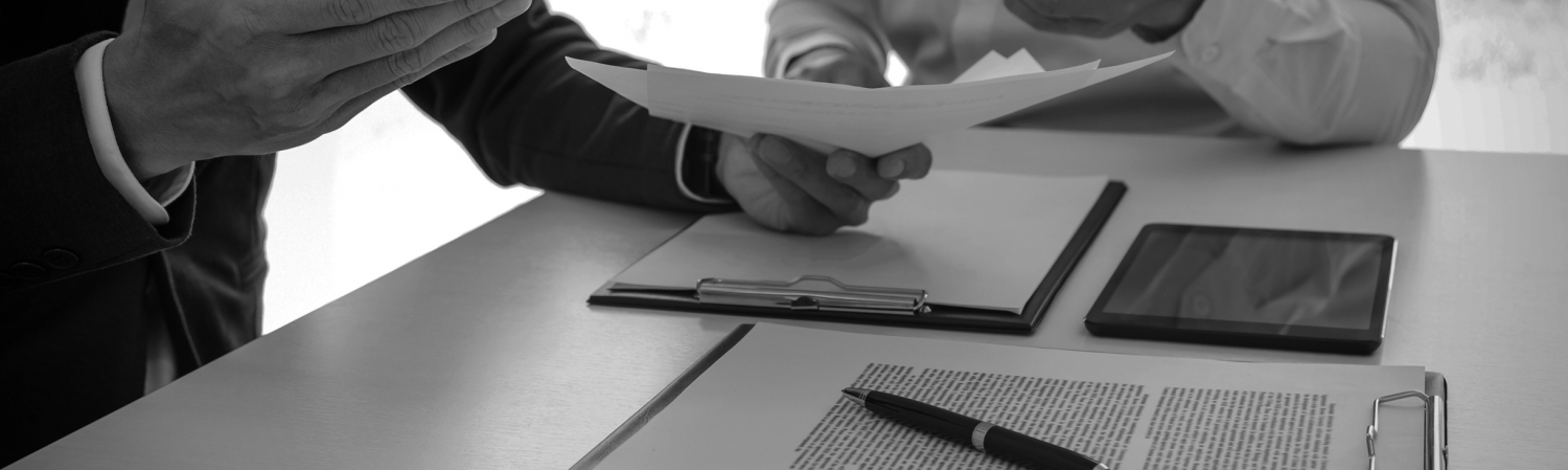 Two men sitting at a table with documents, a tablet, and a calculator discussing a document in one of the man’s hands.