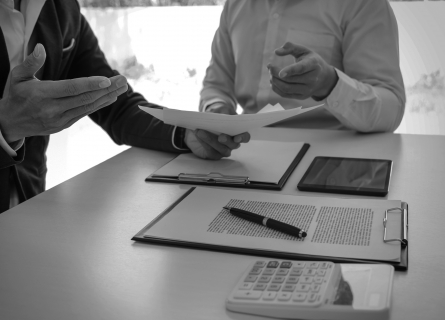 Two men sitting at a table with documents, a tablet, and a calculator discussing a document in one of the man’s hands.