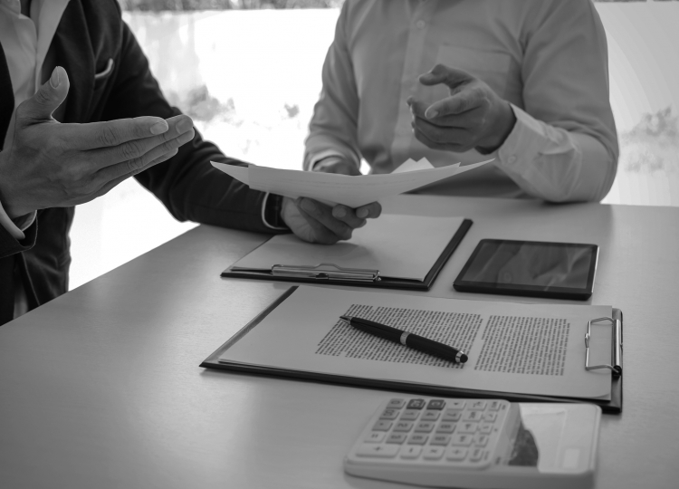 Two men sitting at a table with documents, a tablet, and a calculator discussing a document in one of the man’s hands.