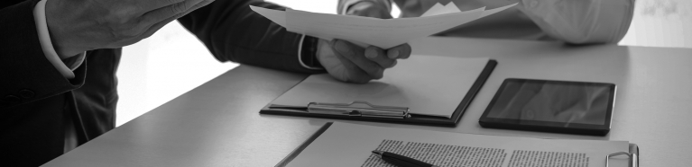 Two men sitting at a table with documents, a tablet, and a calculator discussing a document in one of the man’s hands.