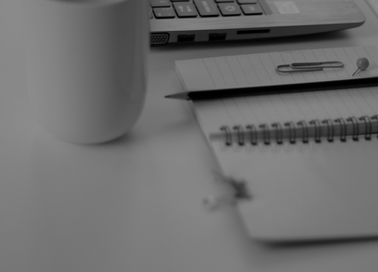 Notebook with a thumbtack, paper clip, and pen on it next to a mug and laptop.