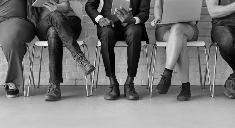 Diverse people sitting in a row of chairs looking at tablets and laptops.