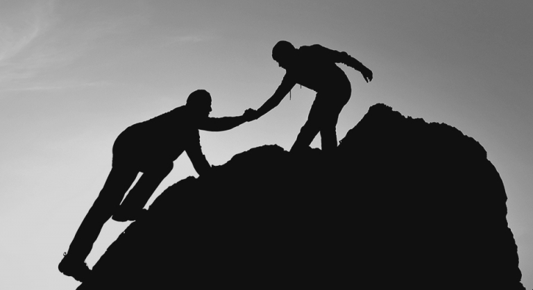 Silhouette of one person helping another reach the top of a rocky mountain.