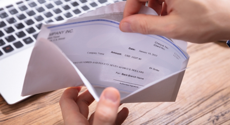 Elevated view of person's hand removing paycheck from the envelope over wooden desk.