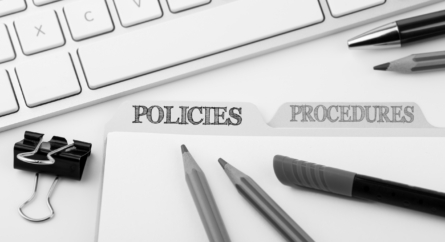 Policies and procedures folders on a desk with a keyboard, paperclips, pens and pencils