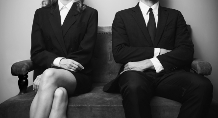 Man and woman dressed in suits sitting on a loveseat with their arms crossed.